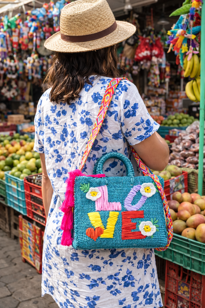 Model wearing a Teal woven LOVE tote bag handmade in Mexico with bright raffia letters and floral accents