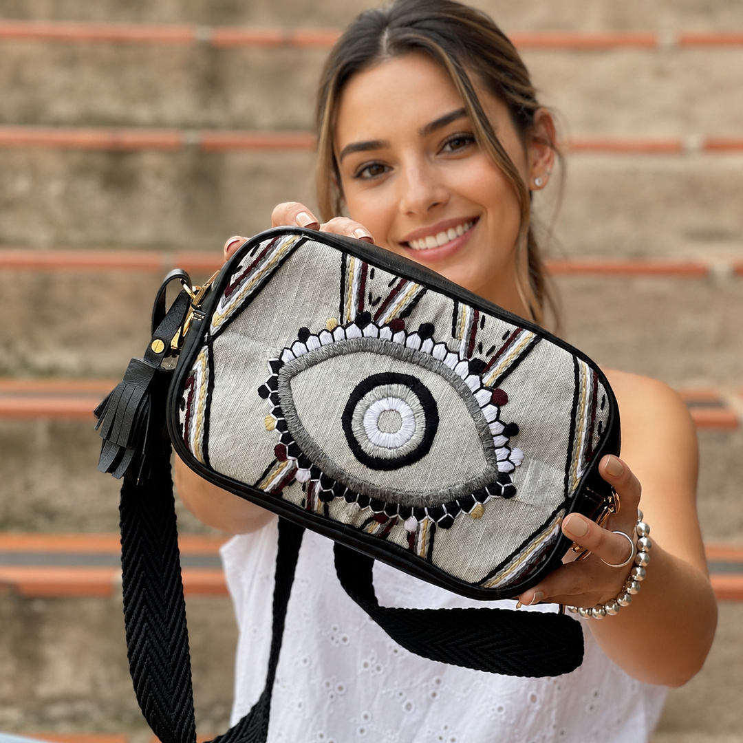 Woman holding embroidered leather evil eye crossbody bag toward the camera outdoors